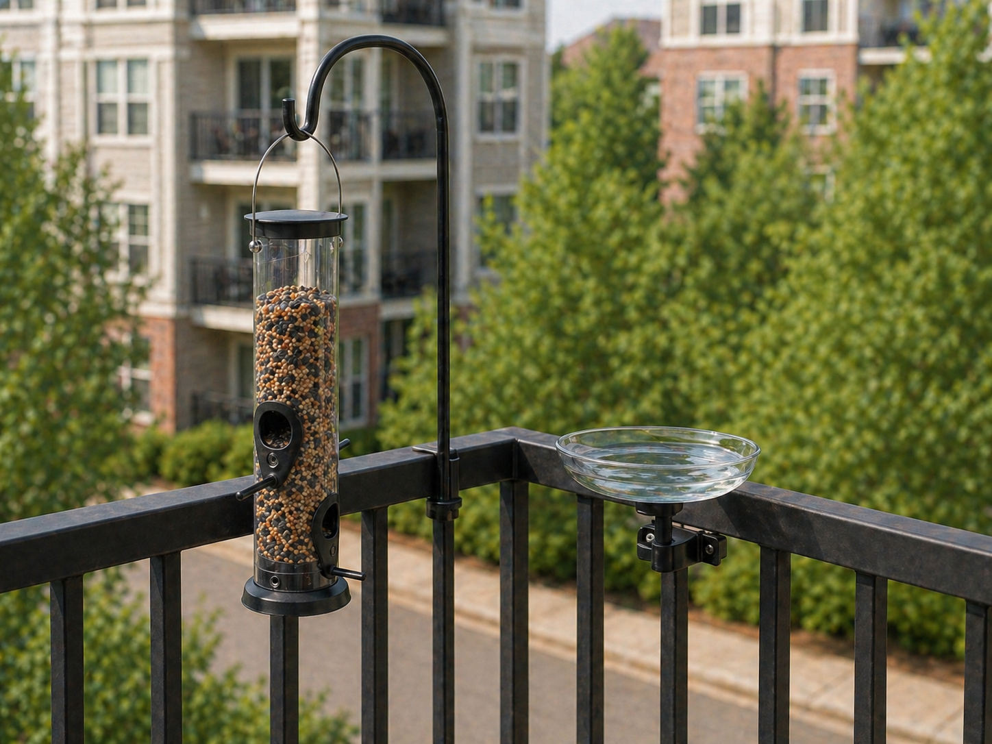 A tube bird feeder hangs from a single shepherd’s hook mounted to a balcony corner, with a shallow clamp-on birdbath attached to the railing.