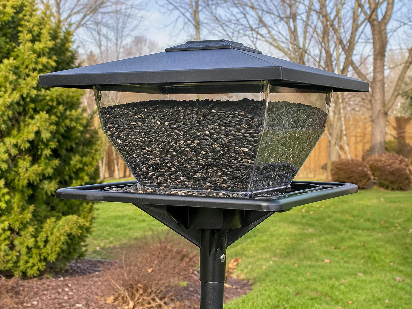 A large black hopper bird feeder filled with black oil sunflower seed is mounted on a pole in a suburban backyard with grass, shrubs, trees, and a wooden fence.